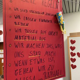 Ein rotes Plakat, auf dem Handschriftlich geschrieben ist: Unsere Regeln! Wir sind freundlich zueinander, wir lassen einander ausreden, wir passen auf unser material auf, wir machen das, was uns Spaß macht, wenn etwas ist, gehen wir zu Alissa und Raphael