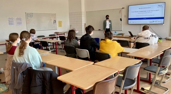 7 Schüler_innen sitzen in einem Klassenraum und schauen auf einen Bildschirm auf dem eine Präsentation angezeigt wird. Zusehen auf der Präsentation ist der Schriftzug "Herzlich Willkommen! Soziale Berufe / Soziale Arbeit. Ist das was für mich?!. 